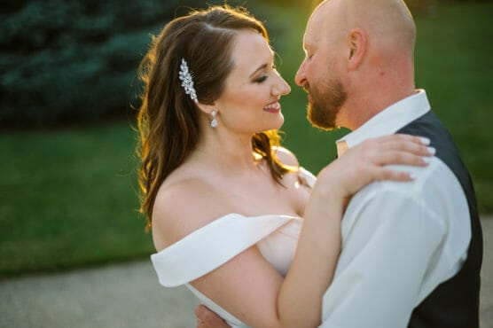 A bride and groom embrace outdoors at sunset, smiling and gazing into each others eyes. The bride wears an off-the-shoulder white dress and a hair accessory, while the groom wears a white shirt and dark vest. by Justin Salem Meyer