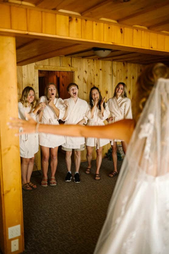 A bride in a wedding dress and veil stands with arms outstretched facing five friends in matching robes, who react with joy and excitement inside a wood-paneled room. by Justin Salem Meyer