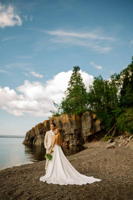 A bride and groom stand on a rocky beach in front of a cliff, with trees and water in the background. The bride wears a long white dress and holds a bouquet; the groom wears a light suit. The sky is partly cloudy. by Justin Salem Meyer