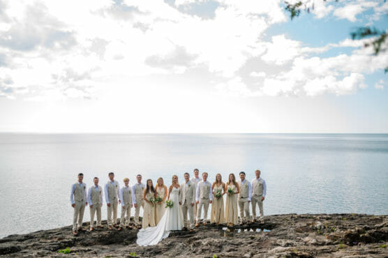 A wedding party stands on rocky ground by a calm, expansive lake under a bright, partly cloudy sky. The group includes people in light-colored attire, with the bride and bridesmaids holding bouquets. by Justin Salem Meyer
