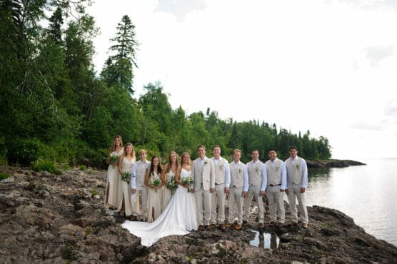 A wedding party in light-colored attire stands on rocky terrain by a calm lakeshore, surrounded by lush green trees and a bright, cloudy sky. The bride and groom are at the center, holding hands. by Justin Salem Meyer