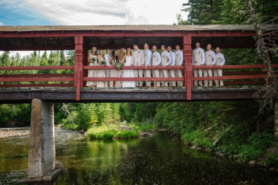 A wedding party poses on a covered red wooden bridge over a river, surrounded by lush green trees and nature on a bright, sunny day. by Justin Salem Meyer