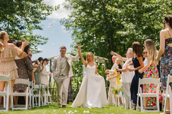 A bride and groom walk hand-in-hand down an outdoor aisle, smiling joyfully as guests on both sides cheer and throw flower petals amid sunny, green surroundings. by Justin Salem Meyer