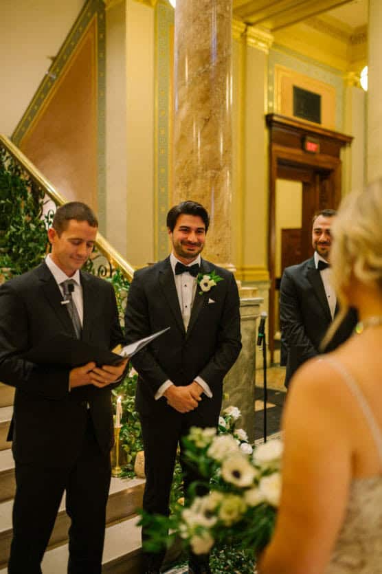 A groom in a black tuxedo smiles warmly at his bride during a wedding ceremony, standing beside an officiant holding a book, with elegant marble columns and greenery in the background. by Justin Salem Meyer