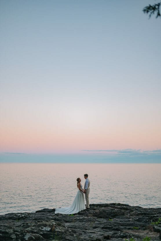 A couple in wedding attire stands on rocky ground by a calm lake at sunset, holding hands under a pastel sky with soft pink and blue hues. by Justin Salem Meyer