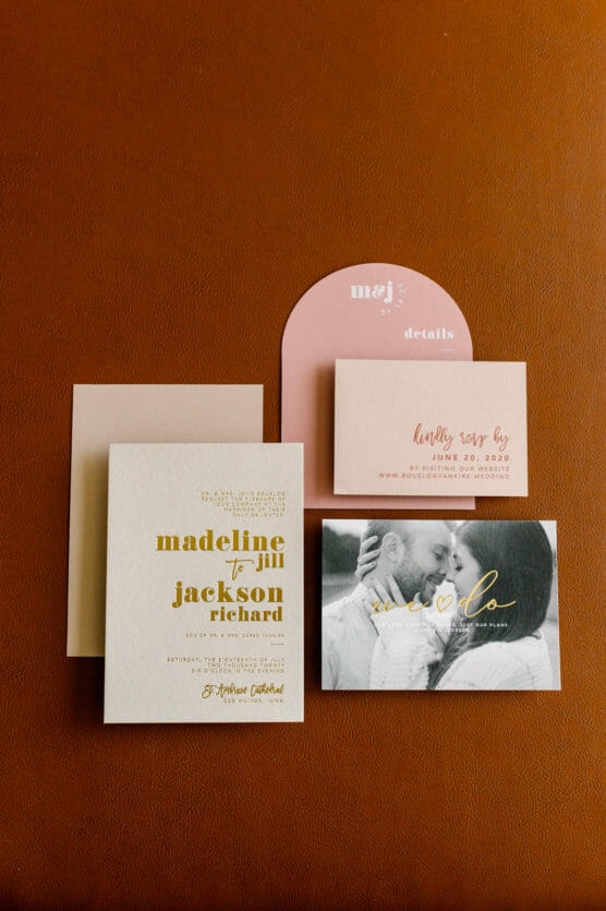 A wedding invitation suite with gold and pink details, including an invitation, RSVP card, details card, and a photo of a couple embracing, all arranged on a brown leather surface. by Justin Salem Meyer