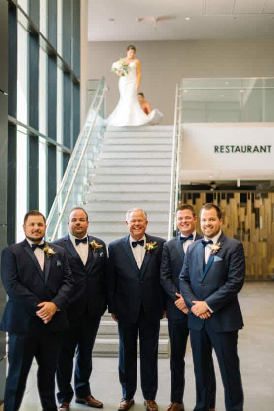 Five men in suits stand together in the foreground, smiling, while a bride in a white gown holds a bouquet and stands at the top of a staircase in the background inside a modern building. by Justin Salem Meyer