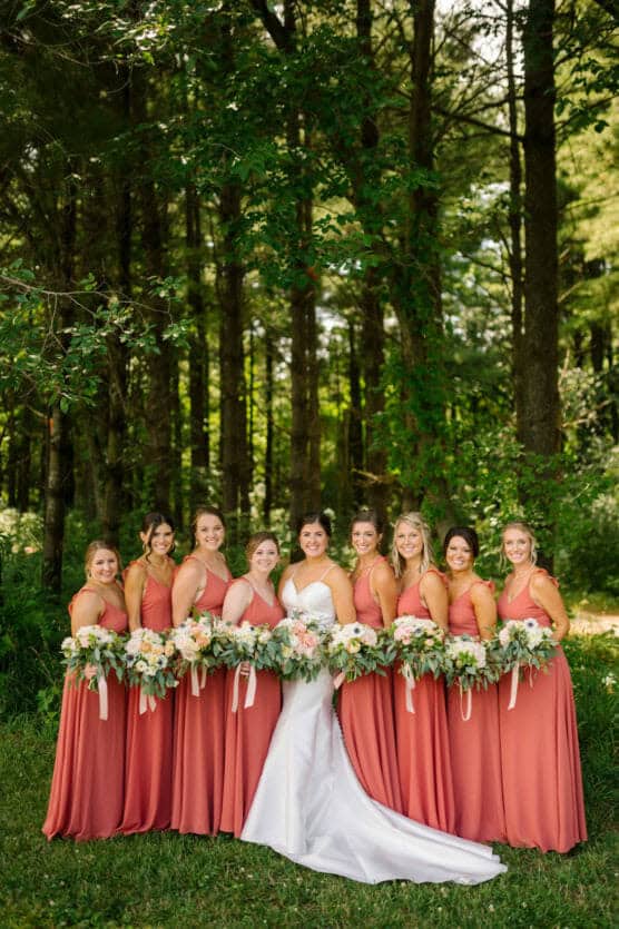 A bride in a white dress stands outdoors with eight bridesmaids in matching rust-colored dresses, all holding bouquets, in front of tall green trees. by Justin Salem Meyer