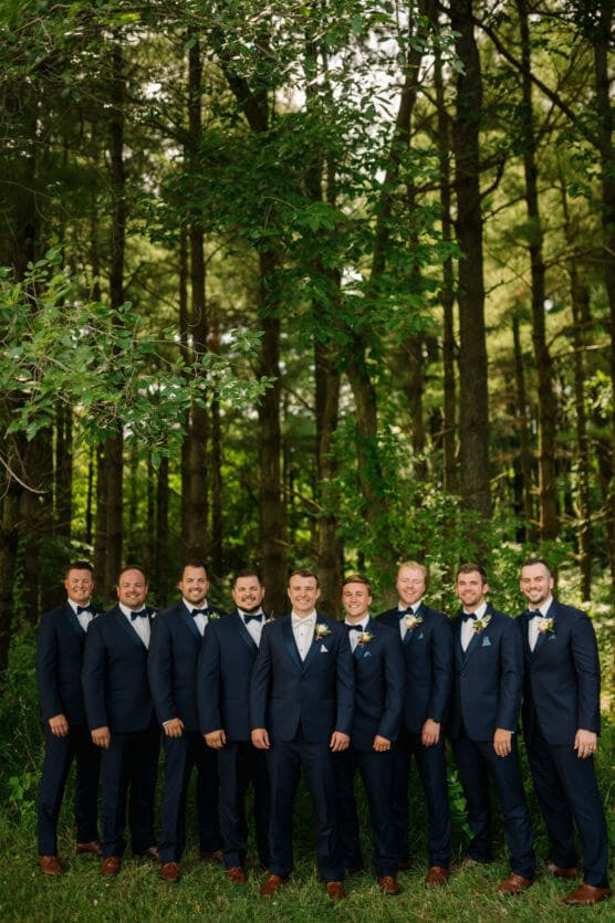 Ten men in matching navy suits and brown shoes stand in a line smiling, posing for a photo in a lush, green forest with tall trees and sunlight filtering through the leaves. by Justin Salem Meyer