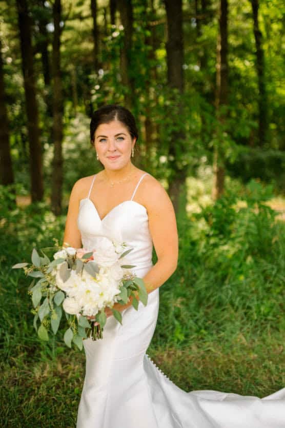 A bride in a white gown holds a bouquet of white flowers and greenery, standing outdoors on grass with a forested background, smiling softly at the camera. by Justin Salem Meyer