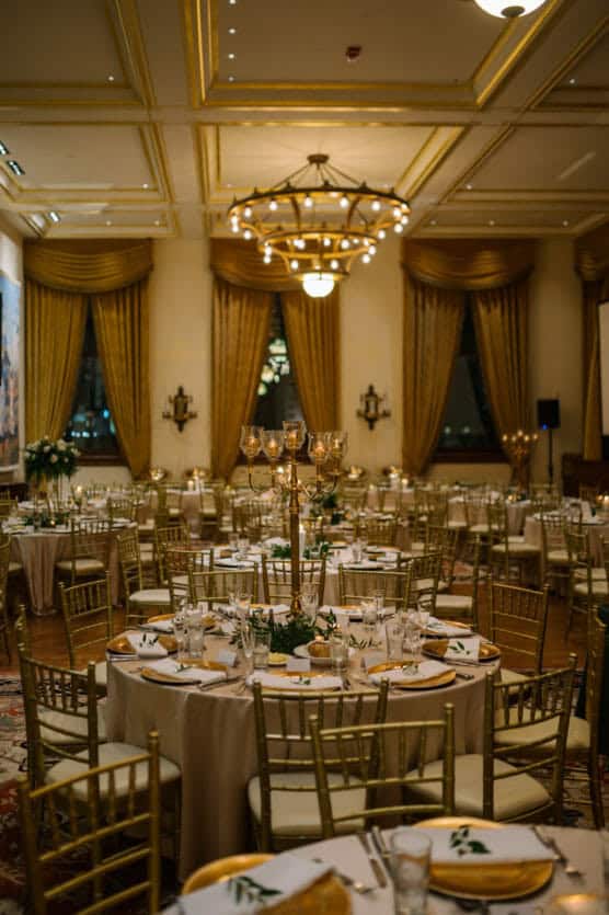 Elegant banquet hall with round tables set for an event, featuring gold chairs, candelabra centerpieces, white tablecloths, and golden drapes. Warm lighting creates a sophisticated, inviting atmosphere. by Justin Salem Meyer