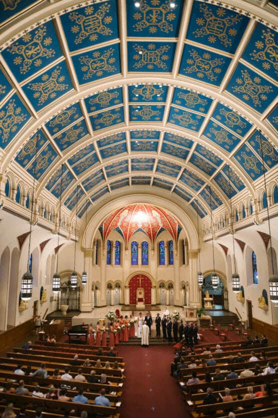 A wedding ceremony takes place in a large church with a blue, gold, and red ornate arched ceiling. Guests are seated in pews, and the wedding party stands at the altar near the officiant. by Justin Salem Meyer