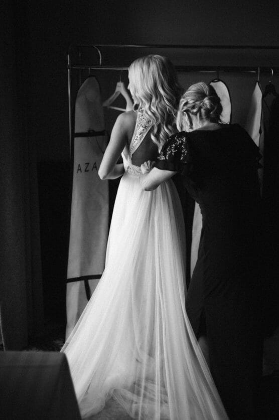 A woman in a wedding dress stands as another woman helps fasten the back of her gown. They are indoors, with garment bags hanging in the background. The scene is in black and white. by Justin Salem Meyer