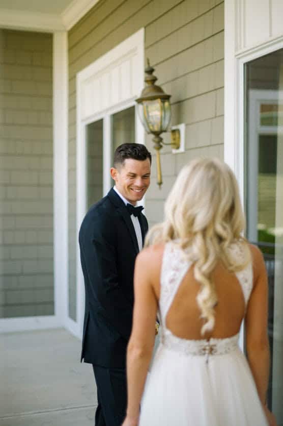 A groom in a black tuxedo smiles at a bride with long blonde hair wearing a white, open-back dress as they stand outside near a building with green siding and large windows. by Justin Salem Meyer