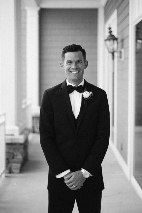 A smiling man in a tuxedo with a bow tie and boutonnière stands on a covered outdoor walkway, posing for a formal portrait; the photo is in black and white. by Justin Salem Meyer