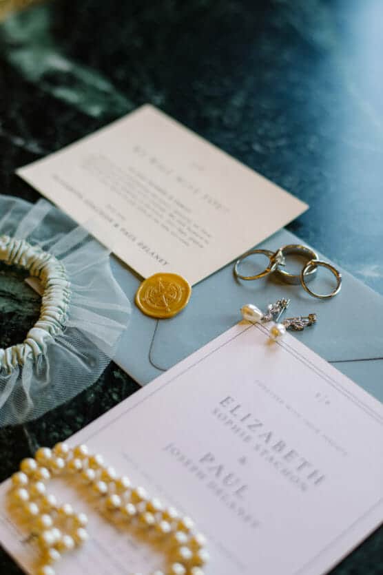 A flat lay of wedding details including invitations, a gold wax seal, a pearl necklace, pearl earrings, two gold rings, a white scrunchie with tulle, and a light blue envelope on a dark marble surface. by Justin Salem Meyer