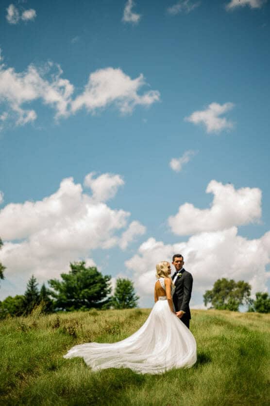 A bride in a flowing white dress and a groom in a suit stand together in a grassy field under a bright blue sky with scattered clouds. by Justin Salem Meyer