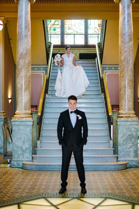 A bride in a white gown stands at the top of a grand staircase holding a bouquet, while a groom in a black tuxedo stands at the bottom with his hands clasped, both in an elegant building with marble columns. by Justin Salem Meyer