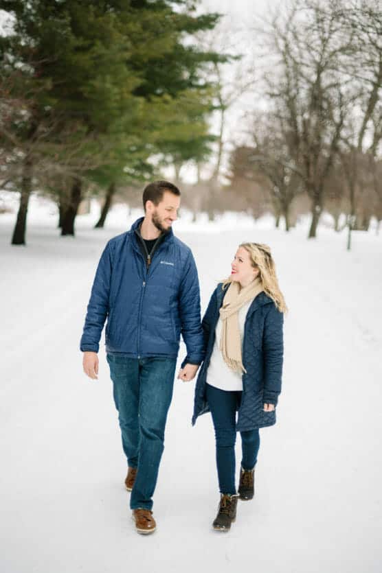 A couple walks hand in hand down a snowy path lined with trees. Both are dressed in winter jackets and boots, smiling at each other as they enjoy the winter scenery. by Justin Salem Meyer