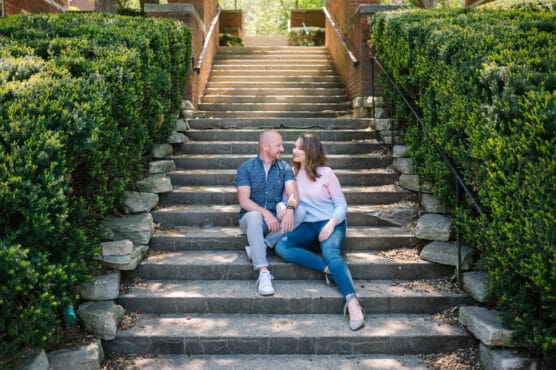 A couple sits together on outdoor stone steps surrounded by green bushes, smiling and looking at each other on a sunny day. by Justin Salem Meyer