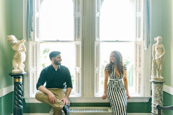 A man and woman sit on a windowsill in a bright room, smiling at each other. Marble statues stand on columns on either side of them. Sunlight streams through tall arched windows with open shutters behind them. by Justin Salem Meyer