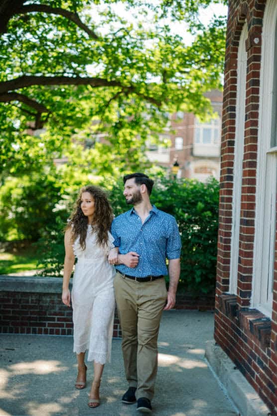 A couple walks arm in arm on a sunny day beside a brick building, surrounded by green trees. The woman wears a white lace dress and heels, while the man wears a blue patterned shirt and khaki pants. They look at each other, smiling. by Justin Salem Meyer
