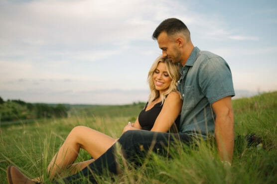 A couple sits closely together in a grassy field at sunset. The woman, smiling, leans against the man, who looks down affectionately at her. The sky is blue with soft clouds, creating a peaceful outdoor scene. by Justin Salem Meyer