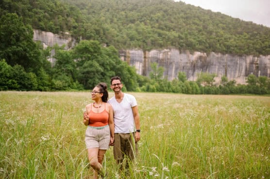 A smiling couple stands in a grassy field with trees and a rocky cliff in the background. The woman wears a coral tank top and shorts, while the man wears a white t-shirt and khaki shorts. by Justin Salem Meyer