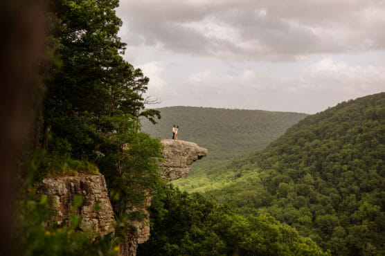A couple stands close together on a rocky cliff edge, surrounded by lush green hills and trees, under a cloudy sky. by Justin Salem Meyer