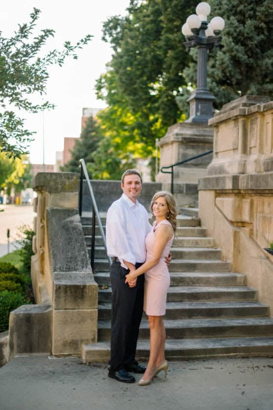 A couple stands on a stone staircase outdoors, holding hands and smiling. The man wears a white shirt and black pants; the woman wears a light pink dress and heels. Trees and a vintage streetlamp are in the background. by Justin Salem Meyer