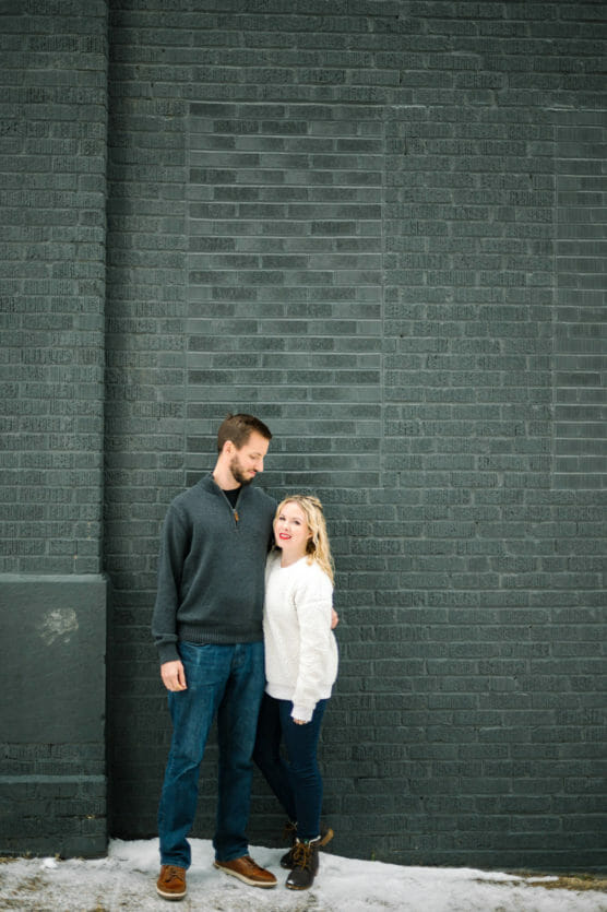 A man and woman stand close together in front of a dark gray brick wall. The man looks down at the woman, who is looking up at him. Both wear sweaters and jeans, with snow on the ground below them. by Justin Salem Meyer