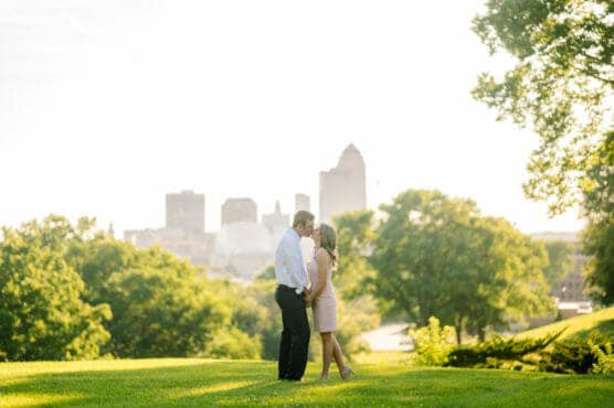 A couple stands close together on a grassy hill, surrounded by trees, with a city skyline in the background under bright, sunny skies. by Justin Salem Meyer