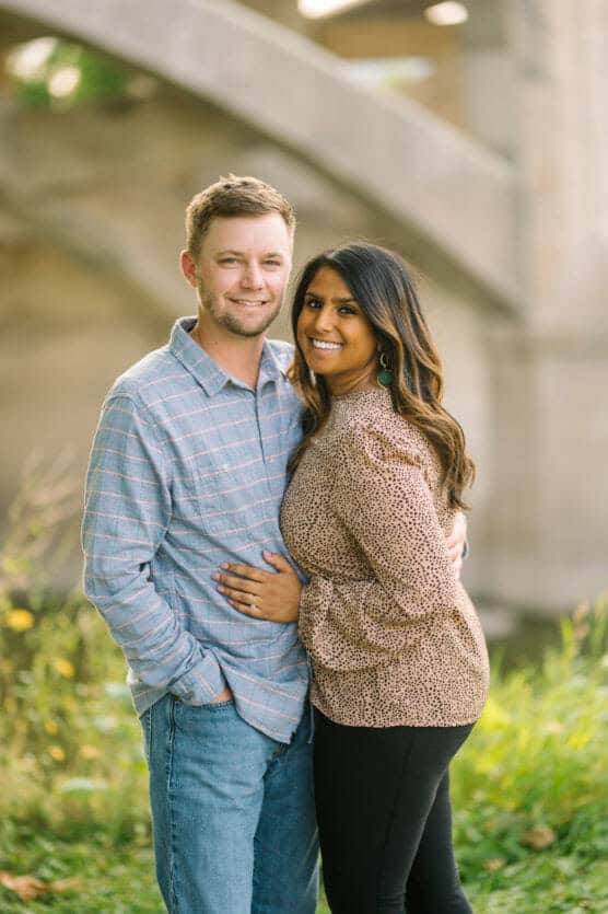 A couple stands outdoors, smiling and embracing. The man wears a light blue plaid shirt and jeans, while the woman wears a leopard-print top and black pants. Blurred greenery and a bridge are in the background. by Justin Salem Meyer