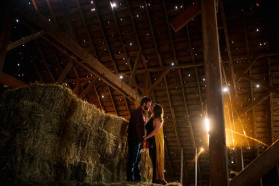 A couple stands on hay bales inside a rustic barn, bathed in warm sunlight streaming through gaps in the wooden roof. They face each other closely, creating a romantic atmosphere. by Justin Salem Meyer