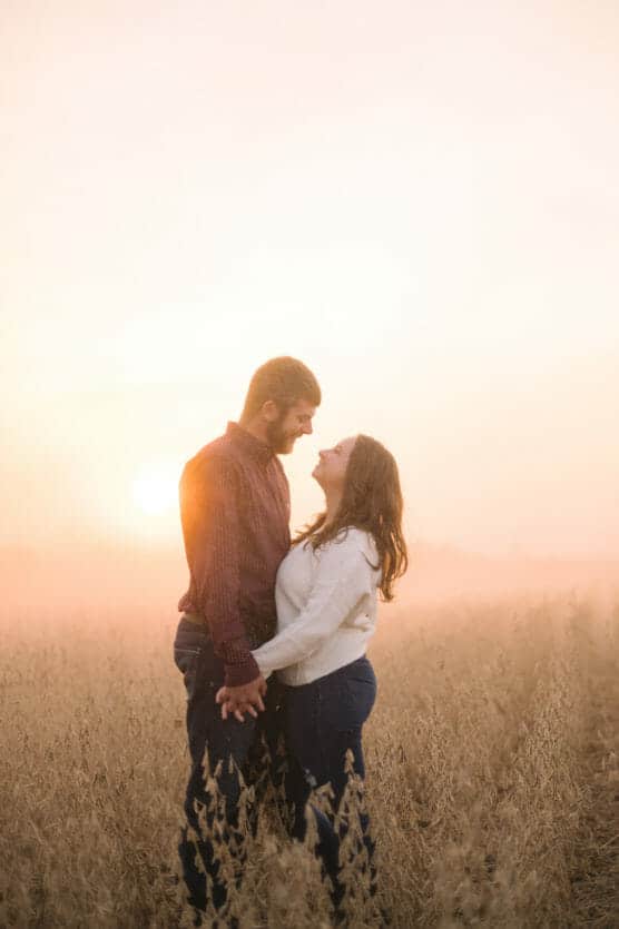 A couple stands close together in a field at sunset, holding hands, gazing into each other’s eyes, with warm sunlight and a soft, hazy sky in the background. by Justin Salem Meyer