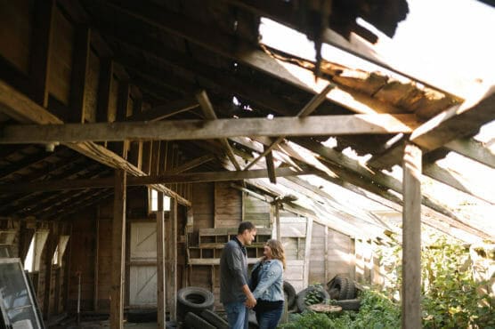 A couple stands holding hands and gazing at each other in an old, partially ruined wooden barn with a broken roof, surrounded by scattered tires and overgrown greenery. by Justin Salem Meyer