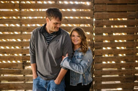 A woman in a denim jacket smiles at the camera while holding the arm of a man in a gray pullover, who looks down with his hands in his pockets. They stand in front of a wooden slat wall with sunlight shining through. by Justin Salem Meyer