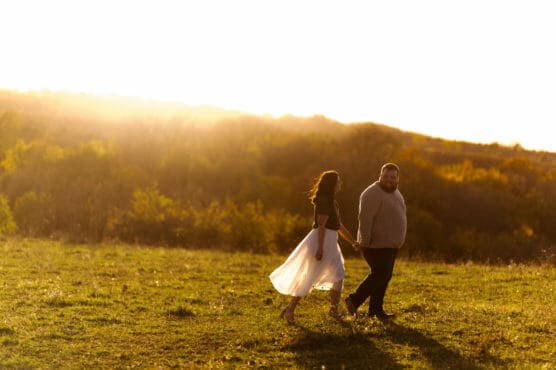 A couple holding hands walks through a grassy field at sunset, with golden sunlight illuminating the landscape and casting a warm glow over the scene. The woman wears a flowing white skirt, and the man wears dark pants and a sweater. by Justin Salem Meyer