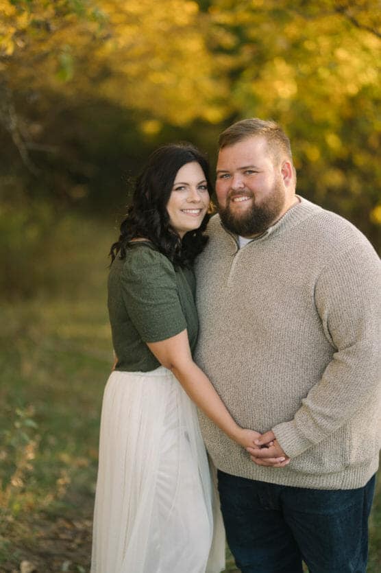 A couple stands close together outdoors, holding hands and smiling at the camera. The woman has dark hair and wears a green top with a white skirt; the man has a beard and wears a beige sweater. Autumn foliage is visible in the background. by Justin Salem Meyer