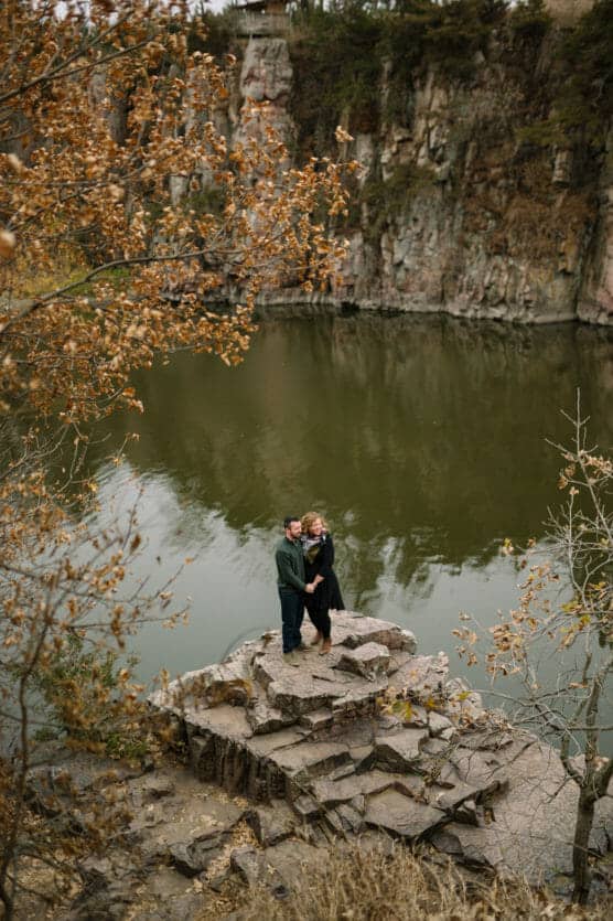A couple stands on a rocky outcrop beside a calm lake, surrounded by autumn trees and cliffs. The water reflects the natural scenery, and fallen leaves are scattered around. The couple appears close and serene. by Justin Salem Meyer