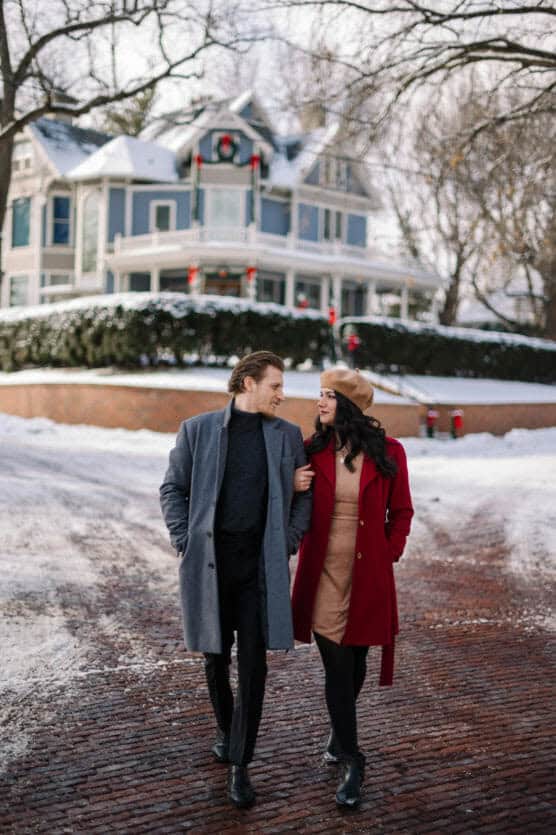 A man and woman walk arm in arm on a snowy street, smiling at each other. They wear winter coats, with the woman in red and the man in gray. Snow-covered Victorian house with holiday wreaths is in the background. by Justin Salem Meyer