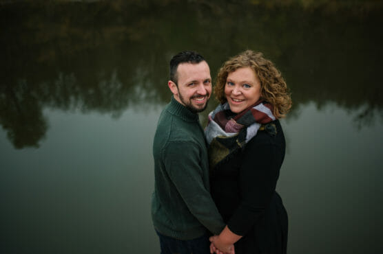 A smiling couple stands close together by a calm body of water, holding hands and looking up at the camera. The man wears a green sweater, and the woman wears a black dress and a multicolored scarf. Trees are reflected in the water behind them. by Justin Salem Meyer