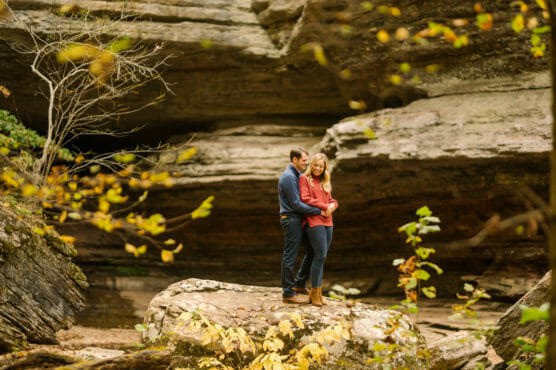 A couple stands close together on a large rock surrounded by autumn foliage and rocky cliffs, sharing an intimate moment in a natural outdoor setting. by Justin Salem Meyer