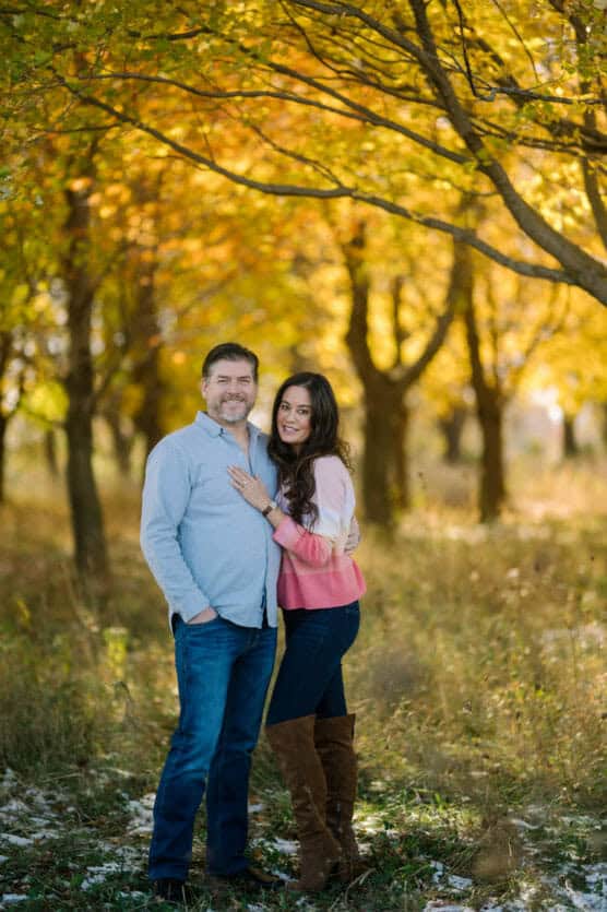 A couple stands smiling together in a sunlit forest with autumn leaves. The man in a light blue shirt and jeans, and the woman in a pink sweater and tall brown boots, are surrounded by golden trees. by Justin Salem Meyer