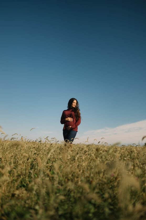 A woman stands in a grassy field under a clear blue sky, wearing a maroon long-sleeve top and dark pants, with long hair flowing and hands resting on her abdomen. by Justin Salem Meyer