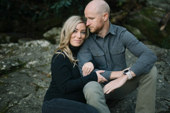A woman with long blonde hair and a man with a shaved head sit close together on rocks outdoors. The woman rests her hand on the mans knee, while he leans in with his arm around her. Both look calm and relaxed. by Justin Salem Meyer
