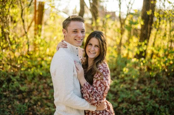 A smiling couple stands close together outdoors, surrounded by green foliage and sunlight filtering through trees. The man wears a light-colored shirt, and the woman wears a maroon dress with white patterns. by Justin Salem Meyer