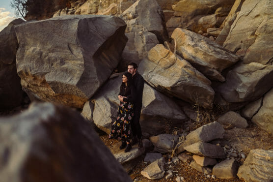 A couple stands close together among large, rugged rocks. The woman wears a black dress with a floral pattern, and the man stands behind her. The lighting is warm, casting soft shadows on the rocky landscape. by Justin Salem Meyer