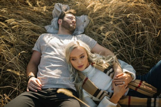 A couple lies in a field of tall, golden grass. The man is resting on his back with his eyes closed, while the woman leans on his chest, holding his arm and looking thoughtfully into the distance. by Justin Salem Meyer