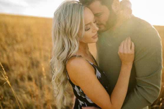 A couple embraces closely in a sunlit field, with tall golden grass surrounding them. The woman has long blonde hair and wears a floral dress, while the man wears a green sweater. They appear affectionate and serene. by Justin Salem Meyer